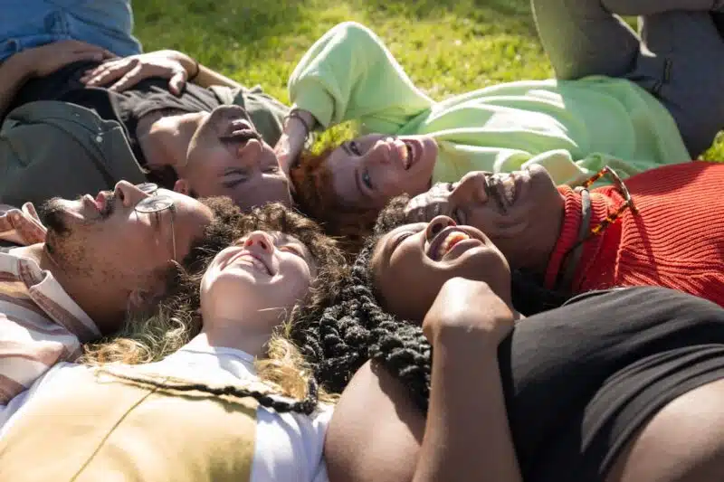 A diverse group of people lying on the grass in a circle, smiling and laughing together, representing connection and belonging in group therapy.