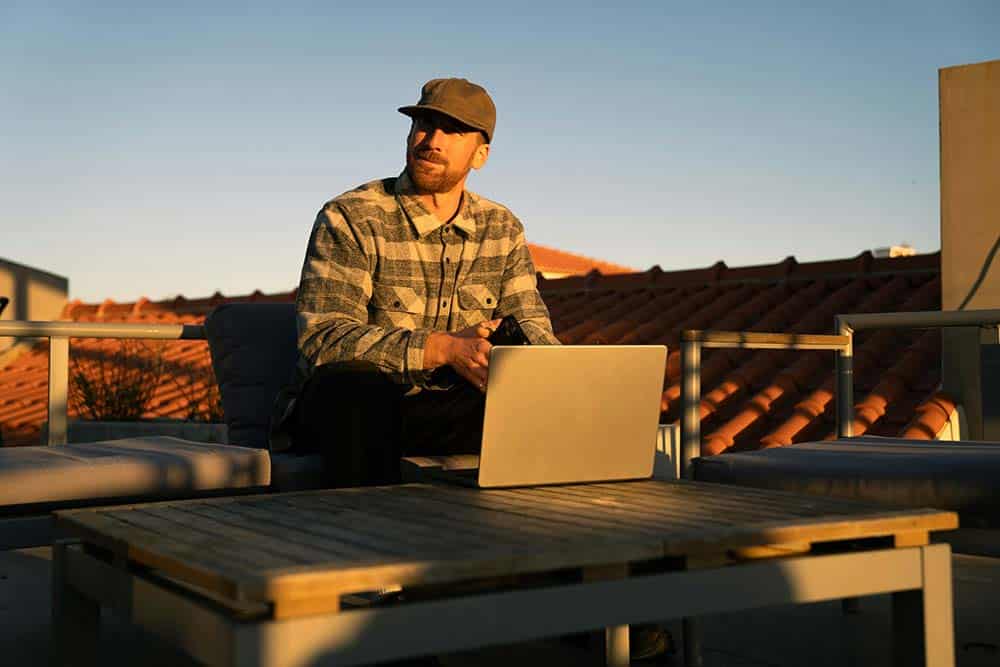 A man in a cap and flannel shirt sitting on a rooftop patio with a laptop and smartphone during sunset.