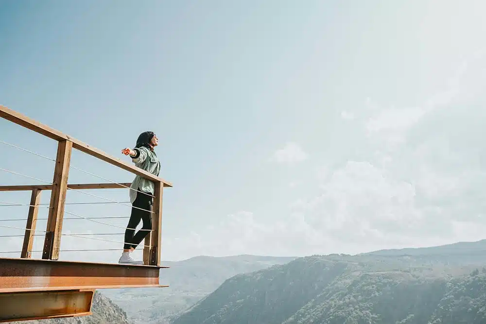 A woman standing on a wooden observation deck with her arms outstretched, overlooking a vast mountain valley under a clear sky.
